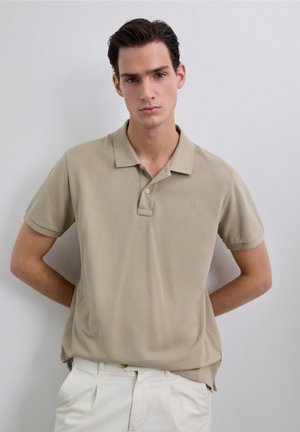 Camisa polo beige con una tela texturizada, mangas cortas, tapeta de tres botones y un sutil bordado de logo en el pecho. Combinada con pantalones blancos.