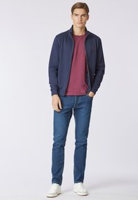 Giacca a zip blu sopra una t-shirt bordeaux, abbinata a jeans blu e sneakers bianche. I tessuti appaiono lisci, con una vestibilità casual e un design semplice.
