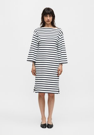 Mujer con cabello oscuro hasta los hombros, vestida con un vestido blanco hasta la rodilla con rayas horizontales negras y zapatos planos negros.