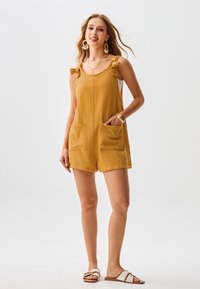 Senfgelber Jumpsuit aus strukturiertem Stoff mit kleinen Rüschenträgern, zwei vorderen Taschen und lockerer Passform. Kombiniert mit flachen Sandalen.