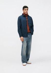 Ναυτικό μπλε σακάκι με κουμπιά, φορεμένο πάνω από ένα σκουροκόκκινο πουλόβερ με λαιμόκοψη, σε συνδυασμό με ανοιχτόχρωμο τζιν φαρδιάς γραμμής και λευκά αθλητικά παπούτσια.