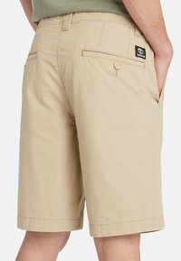 Pantalones cortos de algodón beige con una textura suave, que cuentan con bolsillos laterales y traseros, y un parche con el logo cerca del bolsillo derecho.