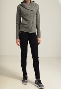 Šedá mikina na zip s asymetrickým límcem, spárovaná s černými úzkými džíny a vzorovanými černými teniskami. Hladká textura, moderní design.