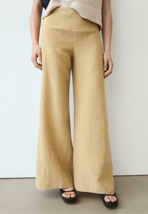 Indossa pantaloni beige a gamba larga con sottili righe verticali, abbinati a sandali platform neri con punta aperta su un pavimento neutro.