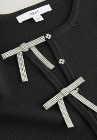Czarny sweter z dzianiny z akcentowanymi kryształkami kokardami i metalowymi zapięciami na zatrzaski. Teksturowany materiał o prążkowanym wzorze. Metka informuje "NEXT."