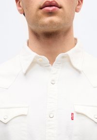 Κάτω μέρος  του προσώπου και λαιμός ατόμου που φοράει λευκό πουκάμισο με κουμπιά και τσέπες στο στήθος, με κόκκινη ετικέτα με το λογότυπο Levi's στην αριστερή πλευρά.