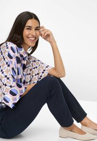 Korte mouw blouse met geometrische patronen in pastelkleuren van roze en blauw, gecombineerd met marineblauwe broek en beige flats.