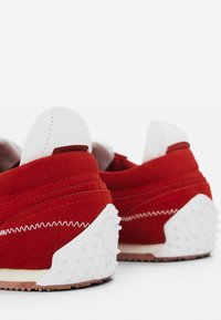 Rote Wildledersneaker mit weißen Akzenten, ausgestattet mit einem gepolsterten Kragen, kontrastierender Naht und einer strukturierten weißen Gummisohle mit kleinen erhabenen Punkten.