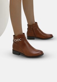Botas de tobillo marrones hechas de cuero suave con un pequeño tacón cuadrado, que presentan un acento decorativo de cadena metálica y cierre de cremallera lateral.