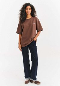 Hnědé oversized tričko s malým bílým vyšívaným detailem, doplněné tmavě modrými rozšířenými džíny a hnědými sportovními botami.