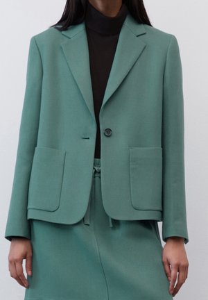 Vrouw met een groene, op maat gemaakte blazer over een zwarte coltrui en bijpassende groene broek met voorzakken en een knoopsluiting.