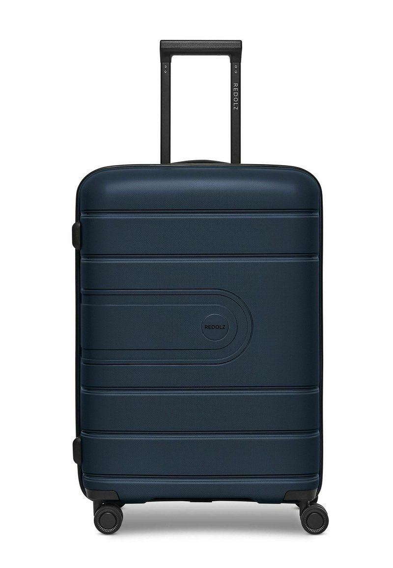 Valigia rigida blu navy con strisce orizzontali in rilievo, manico telescopico nero e quattro ruote nere per la mobilità. Logo sulla parte anteriore.