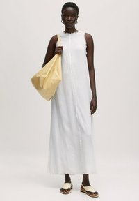 Weißes ärmelloses Maxikleid mit fransigem Saum, kombiniert mit einer gelben Lederhandtasche. Model trägt flache beigefarbene Sandalen und steht vor einem neutralen Hintergrund.