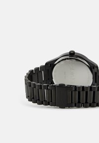 Reloj de metal negro con un diseño elegante, que presenta una correa segmentada y una caja redonda. La parte posterior es lisa y grabada con los detalles de la marca.