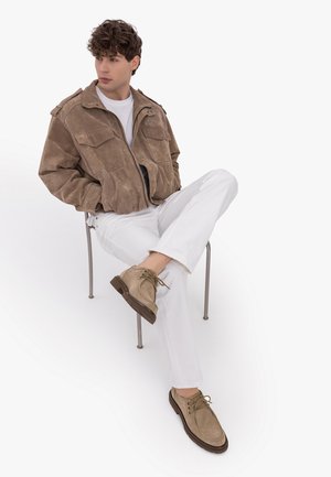 Young man sitting on metal chair, wearing brown suede jacket, white pants, white shirt, and tan lace-up shoes, looking to the side.