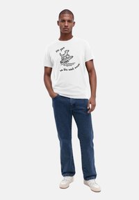 Weißes Baumwoll-T-Shirt mit einem schwarzen Grafik eines Hundes auf einem Surfbrett und dem Text "See you on the next wave", kombiniert mit blauen Jeans.
