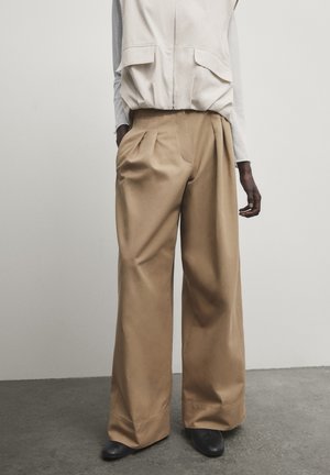 Silhouette élancée portant un pantalon beige à jambes larges, un gilet blanc zippé devant et des chaussures noires, debout sur un sol en béton devant un mur gris uni.