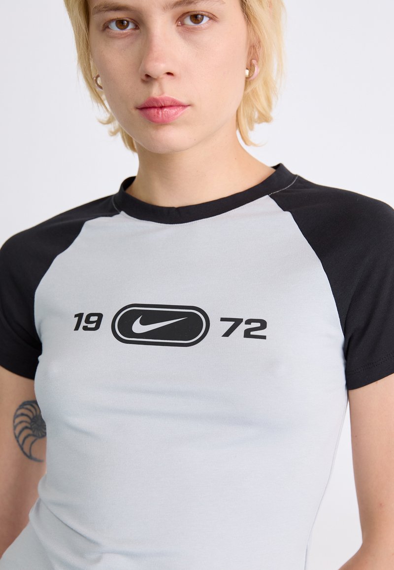 Camiseta de manga corta gris claro con mangas raglán negras, que presenta un logo de Nike negro en el centro y "1972" impreso debajo. Textura de tela suave.