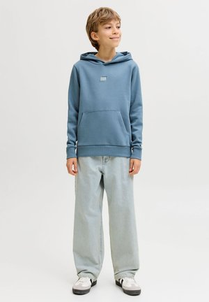 Αγόρι που στέκεται φορώντας μπλε φούτερ με κουκούλα, ανοιχτό μπλε τζιν με φαρδιά πόδια και λευκά αθλητικά, κοιτάζοντας ελαφρώς στο πλάι με ένα χαμόγελο.