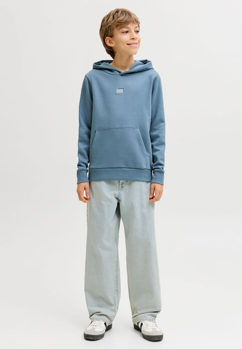 Αγόρι που στέκεται φορώντας μπλε φούτερ με κουκούλα, ανοιχτό μπλε τζιν με φαρδιά πόδια και λευκά αθλητικά, κοιτάζοντας ελαφρώς στο πλάι με ένα χαμόγελο.