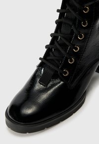 Botín negro hasta el tobillo con un acabado brillante de charol, diseño de cordones, ojales dorados, tejido texturizado en el lateral y un tacón bajo cuadrado.