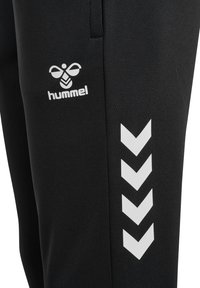 Pantalon de sport noir avec logo Hummel blanc et quatre chevrons pointant vers le bas sur la cuisse gauche.