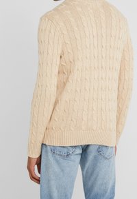 Beżowy sweter w splot kablowy z prążkowanym dołem i mankietami, z charakterystyczną teksturą. Noszony na jasnoniebieskich dżinsach, widok z tyłu.