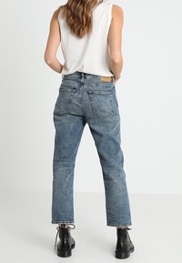 Svetle modré džíny s vysokým pasem a rovnými nohavicemi, se zadními náprsními kapsami a koženou cedulkou. V kombinaci s černými kotníkovými botami.