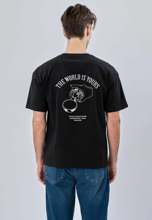Mann trägt ein schwarzes T-Shirt mit weißem Grafikbild einer Hand, die einen Globus hält, und dem Text „DIE WELT GEHÖRT DIR“ und „STIL IST VIEL INTERESSANTER ALS MODE“.