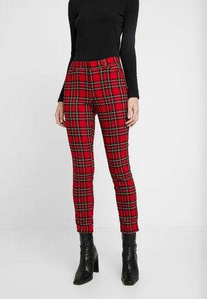 Kvinde iført stramme røde ternede bukser, sort langærmet top og sorte ankelstøvler med hæl mod en ensfarvet baggrund.