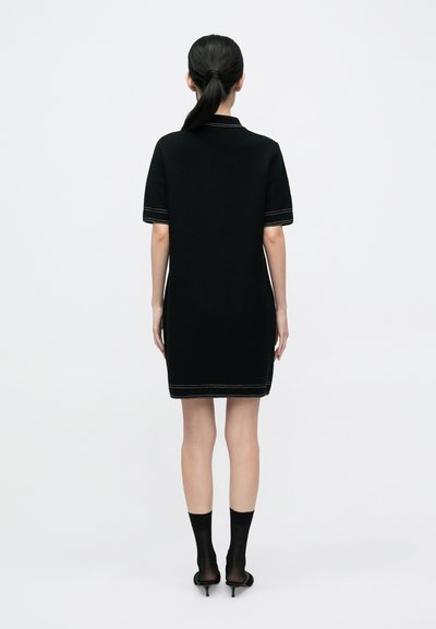 Fekete kötött rövid ujjú pólóruha fehér varrással a szegély mentén. Áttetsző fekete zoknival és hegyes orrú magassarkúval párosítva.