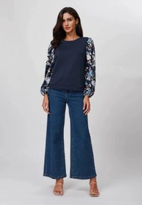 Marinblå hettegenser med blomstermønstrede ballongermer, kombinert med vide blå denimjeans og nøytrale stroppsandaler.