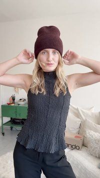 Mlada žena prilagaja bordo pleteno kapo, oblečena v brezrokavni črn top z teksturo in črne hlače, stoji v svetlem dnevnem prostoru z zelenim omarico.