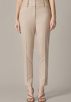 Beżowe, dopasowane spodnie za kostkę z przednimi fałdami, noszone z brązowymi sandałami na wysokim obcasie z paskami na neutralnym tle.