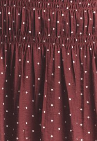 Burgundy-kangas valkoisilla pilkuilla, jossa on kerroksellinen tekstuuri ja joustava vyötärö. Pehmeä, kevyt materiaali, jossa on sileä pinta.