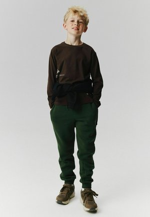 Lyshåret dreng står med hænderne i lommerne iført mørk langærmet skjorte, grønne bukser, brune sneakers og sort sweater bundet om livet.