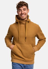 Haut à capuche jaune moutarde avec un motif texturé, cordons de serrage ajustables, poche avant et poignets côtelés. Le mannequin le porte sur un jean bleu.