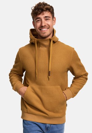 Sudadera color mostaza con patrón texturizado, cordones ajustables, bolsillo frontal y puños acanalados. El modelo la lleva puesta sobre unos vaqueros azules.