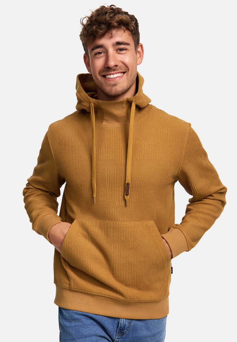 Haut à capuche jaune moutarde avec un motif texturé, cordons de serrage ajustables, poche avant et poignets côtelés. Le mannequin le porte sur un jean bleu.