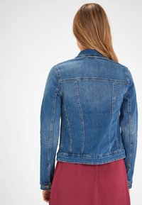 Dopasowana kurtka dżinsowa w średnim odcieniu niebieskiego, z klasycznym kołnierzem, przeszyciami i mankietami na guziki. Teksturowana tkanina z delikatnym efektem wyblaknięcia.