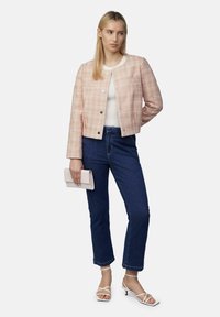 Roze tweed jas met een ronde halslijn en knoopsluiting, gecombineerd met donkerblauwe jeans en witte sandalen met bandjes. Houdt een lichtroze clutch vast.