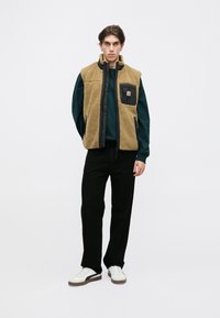 Chaleco de forro polar color beige con ribetes negros y un bolsillo en el pecho, combinado con una camisa de manga larga color verde oscuro y pantalones negros; zapatillas blancas.