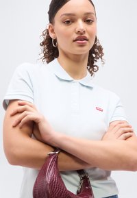 Νεαρή γυναίκα που φοράει ανοιχτό μπλε πόλο μπλούζα με το λογότυπο της Levi's, κρατώντας μπορντό τσάντα με υφή, ποζάροντας με σταυρωμένα τα χέρια μπροστά σε απλό φόντο.