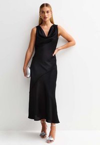 Vestito nero senza maniche con scollo a cowl, tessuto fluttuante, orlo asimmetrico, abbinato a accessori argento e sandali con tacco.