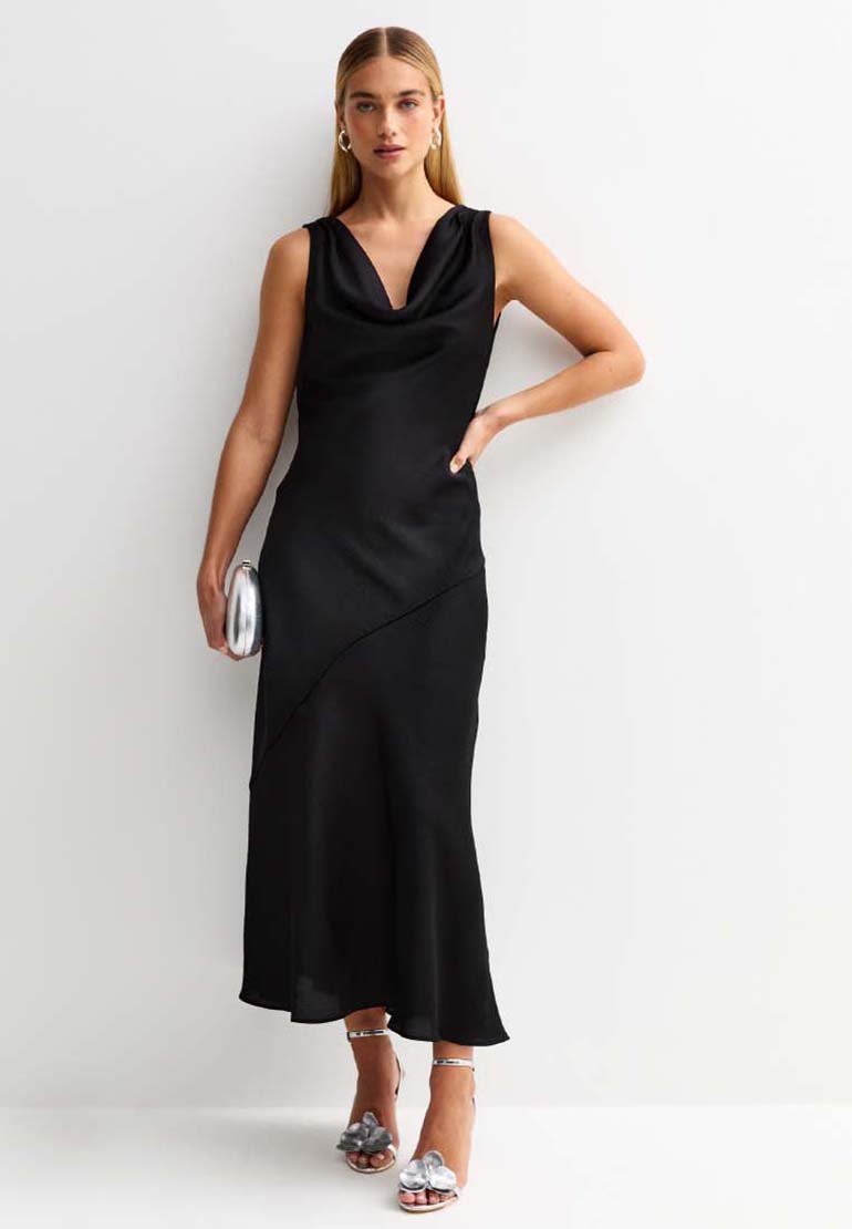 Vestito nero senza maniche con scollo a cowl, tessuto fluttuante, orlo asimmetrico, abbinato a accessori argento e sandali con tacco.