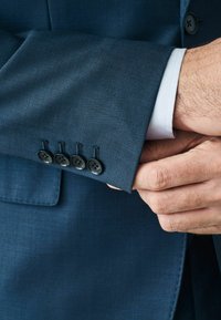 Blaues Sakko mit vier schwarzen Knöpfen am Ärmel, strukturierter Stoff und sichtbarer weißer Hemdmanschette darunter. Maßgeschneiderte Passform.