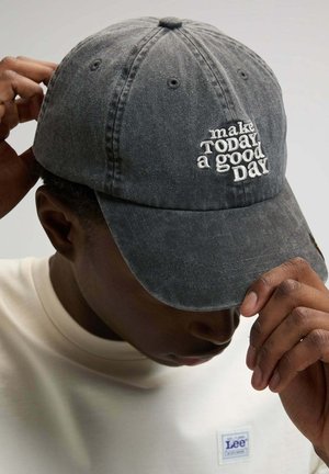 Person trägt eine verblasste schwarze Kappe mit der Stickerei "make TODAY a good DAY", richtet den Kappenschirm aus und trägt ein weißes Hemd der Marke Lee.