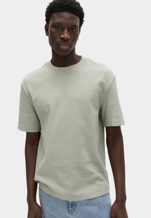 Mann mit dunkler Haut und kurzen lockigen Haaren, der ein hellgrünes, strukturiertes kurzärmeliges Hemd und blaue Jeans vor einem einfarbigen Hintergrund trägt.