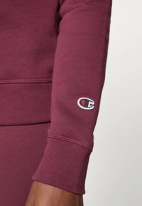 Kastanienbrauner Sweatshirtärmel mit geripptem Bündchen und kleinem gesticktem Champion-Logo am Unterarm, getragen von einer dunkelhäutigen Person.