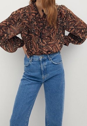 Kobieta w brązowej bluzce w wzór paisley, włożonej do wysokich niebieskich dżinsów, stoi z rękami na biodrach.